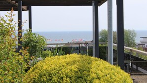 A covered outdoor area with a view of the sea, featuring metal columns, glass railings, and yellow flowering plants. - "Sunny spot" dreamy sunsets included! (Dahme)
