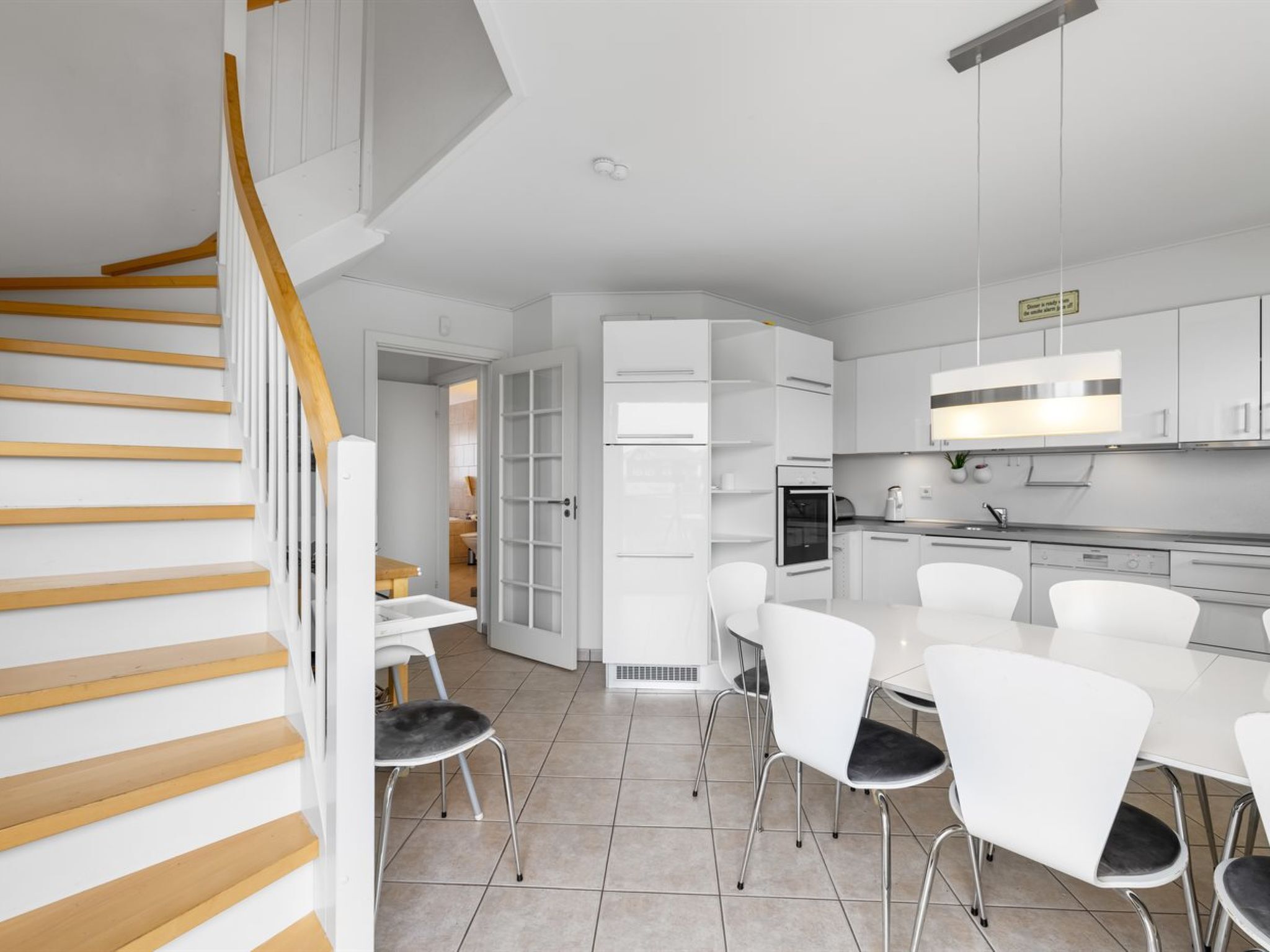 A modern kitchen with white cabinetry, a dining area with white chairs, and a staircase with wooden steps.
