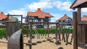 Un terrain de jeux avec des structures en bois et des pneus, le tout installé sur un sol sablonneux, et entouré de bâtiments aux toits rouges.