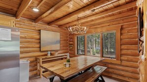 Interior de una cabaña de madera con una mesa, bancos, un refrigerador, un cuadro y ventanas con vista a un bosque.