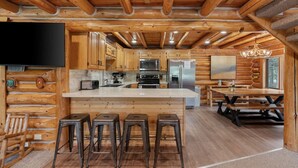 Interior de una cabaña de madera con cocina, taburetes de bar, una mesa de comedor y un televisor.