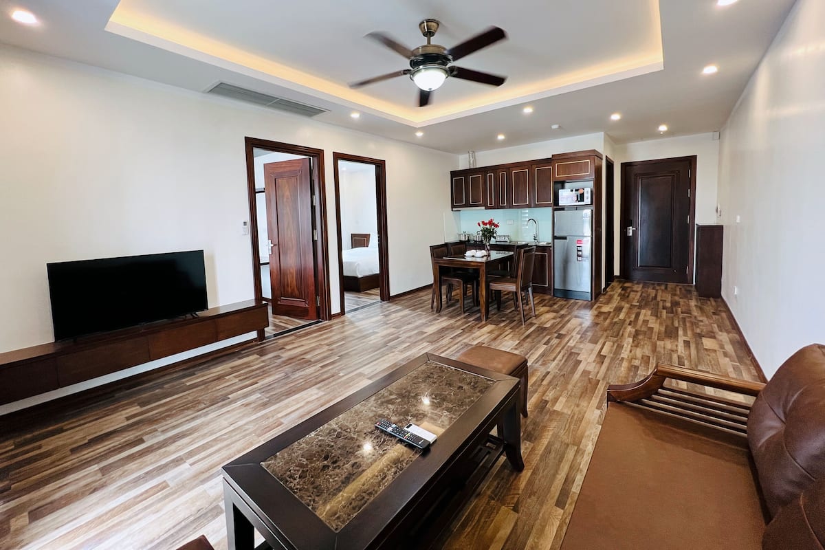 A modern living room with a flat-screen TV, a coffee table, and a sofa. There is a kitchen area with wooden cabinets and a refrigerator.