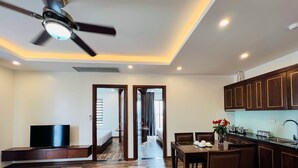 A hotel room with a wooden floor, a TV, a ceiling fan, a dining area with a table and chairs, and a kitchen with wooden cabinets. - Hanoi Apartment  (Hanoi)