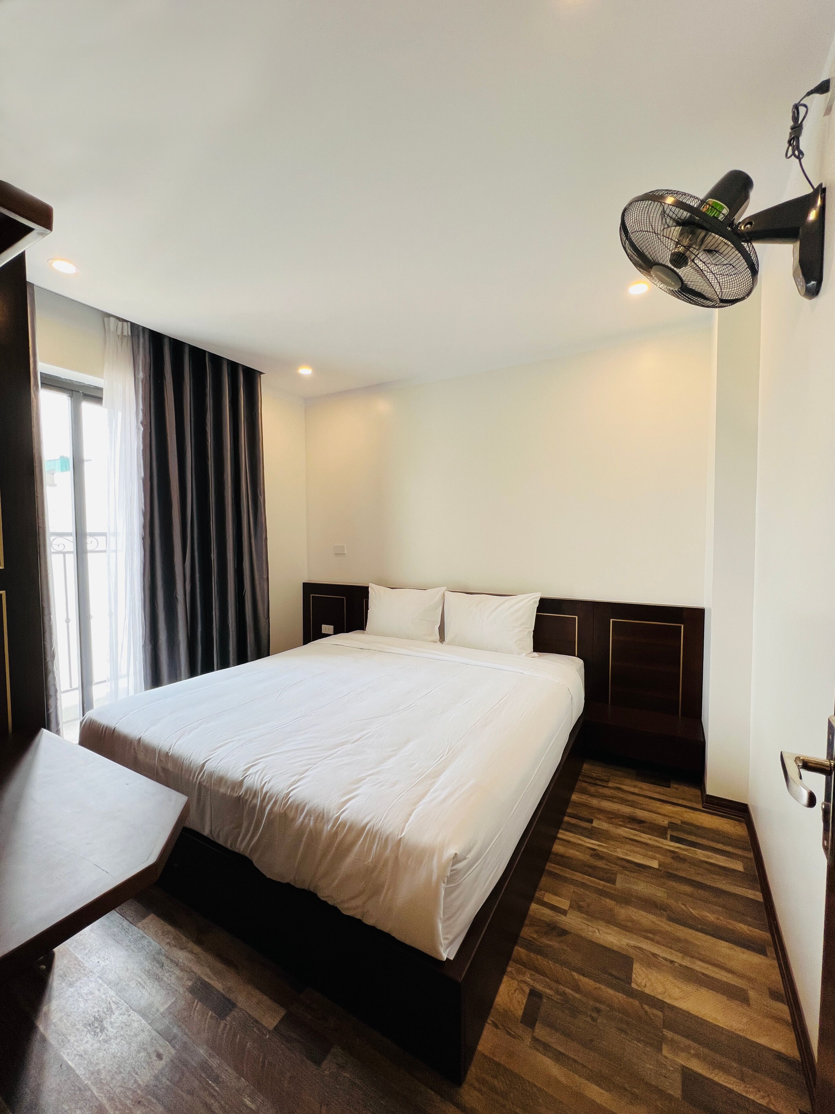 A hotel room with a large bed, a bedside table, a wall-mounted fan, and a view of the outdoors through a window with curtains.