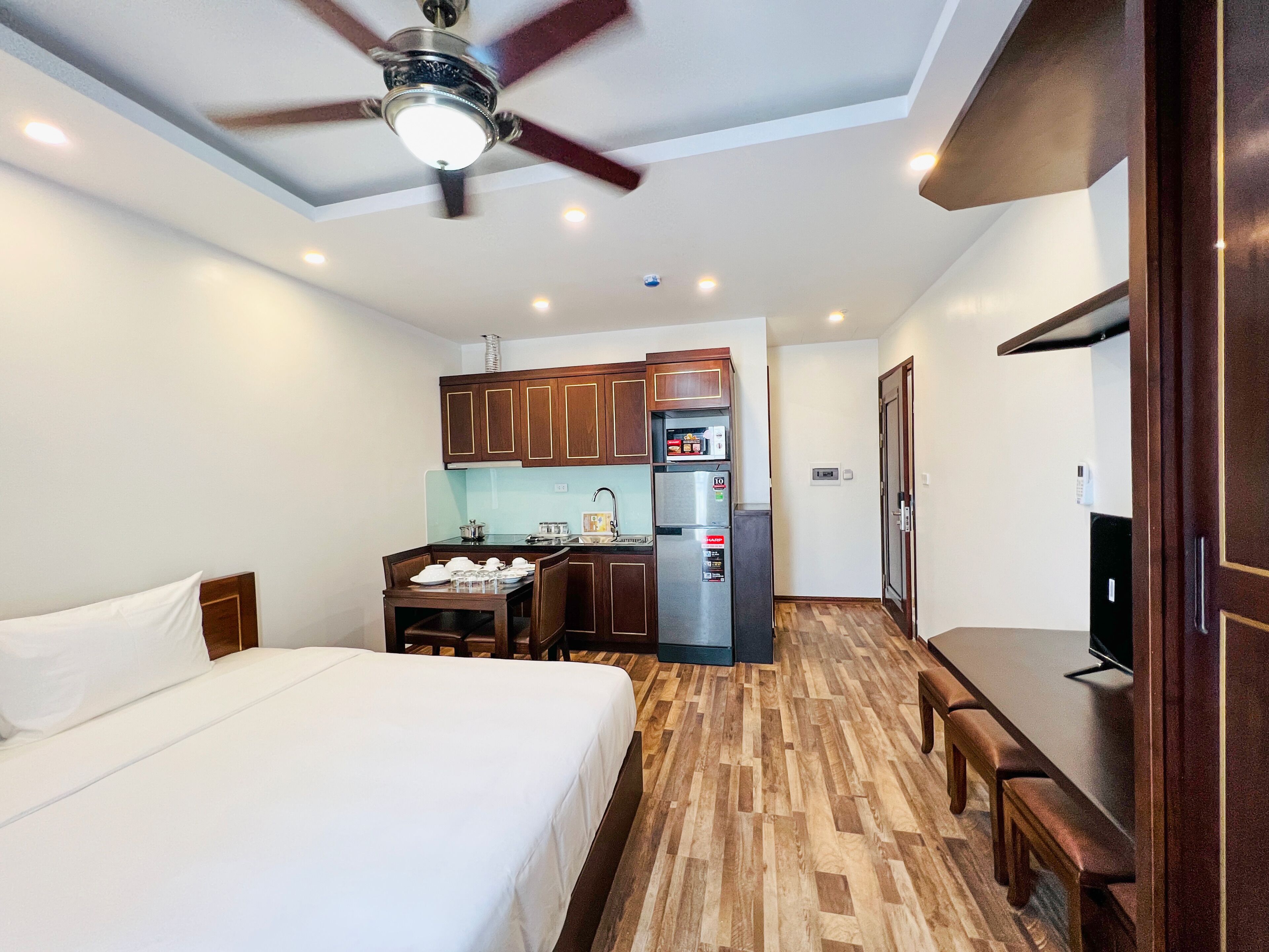 A hotel room with a bed, a desk, a kitchenette, and a ceiling fan.