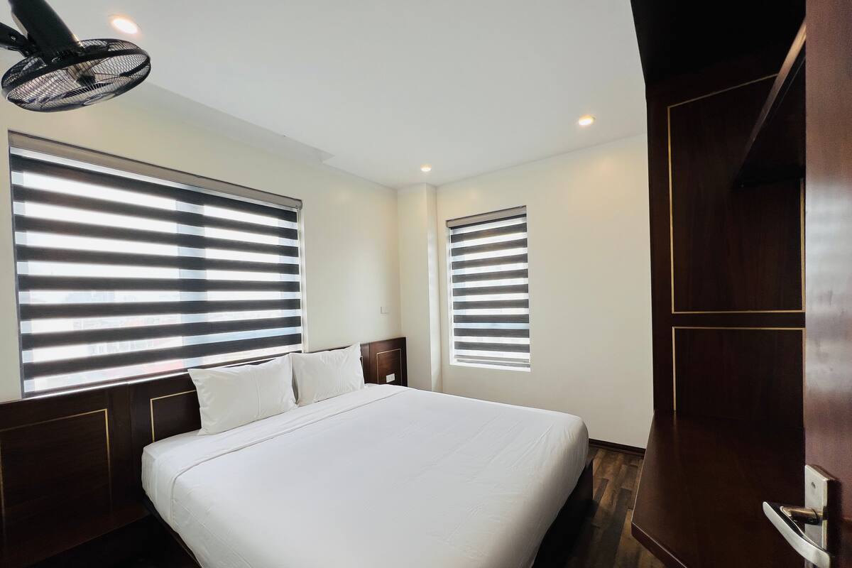 A hotel room with a large bed, two windows with blinds, a ceiling fan, and a wooden wardrobe.