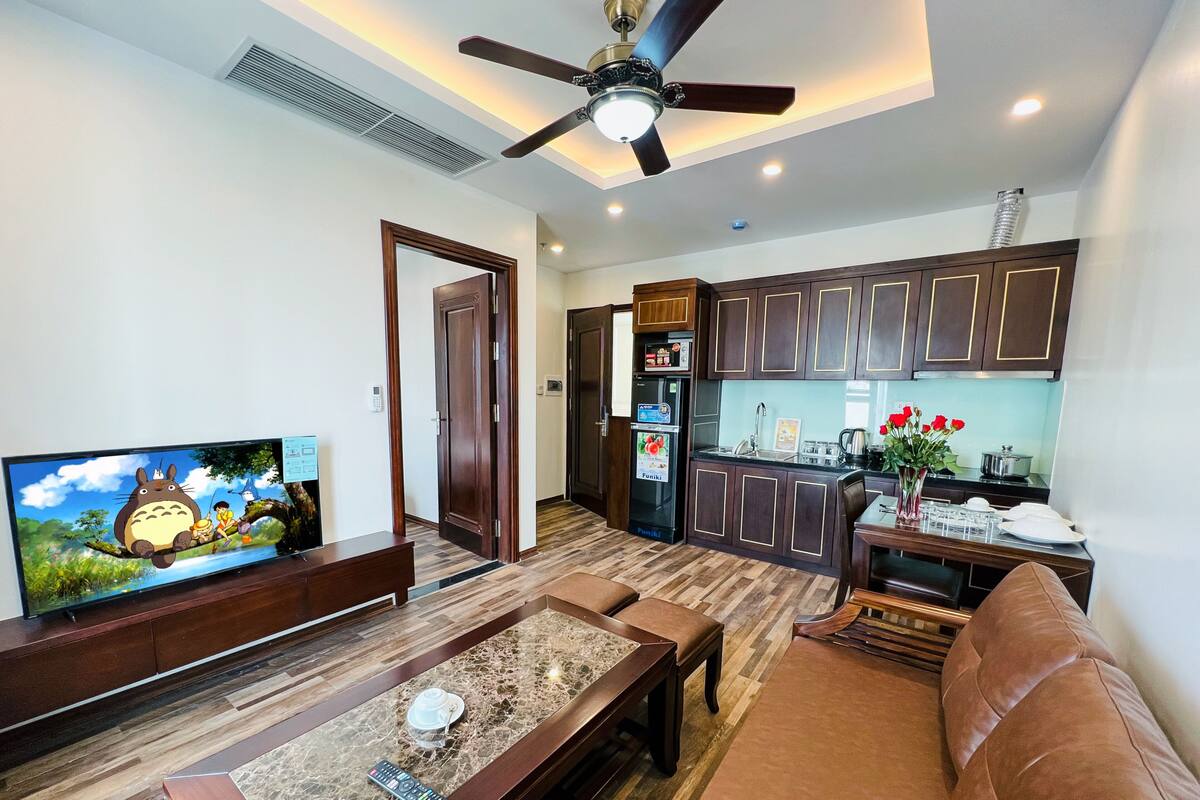 A living room with a TV, sofa, coffee table, and a kitchen area with cabinets and a refrigerator.
