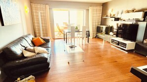 Ein Wohnzimmer mit einer schwarzen Ledersofa, einem Couchtisch und einem Fernseher. Eine Glastür führt auf einen Balkon.