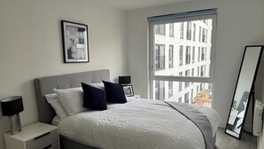 A modern bedroom with a large bed, a nightstand, a bedside table, a mirror, and a window with a city view.