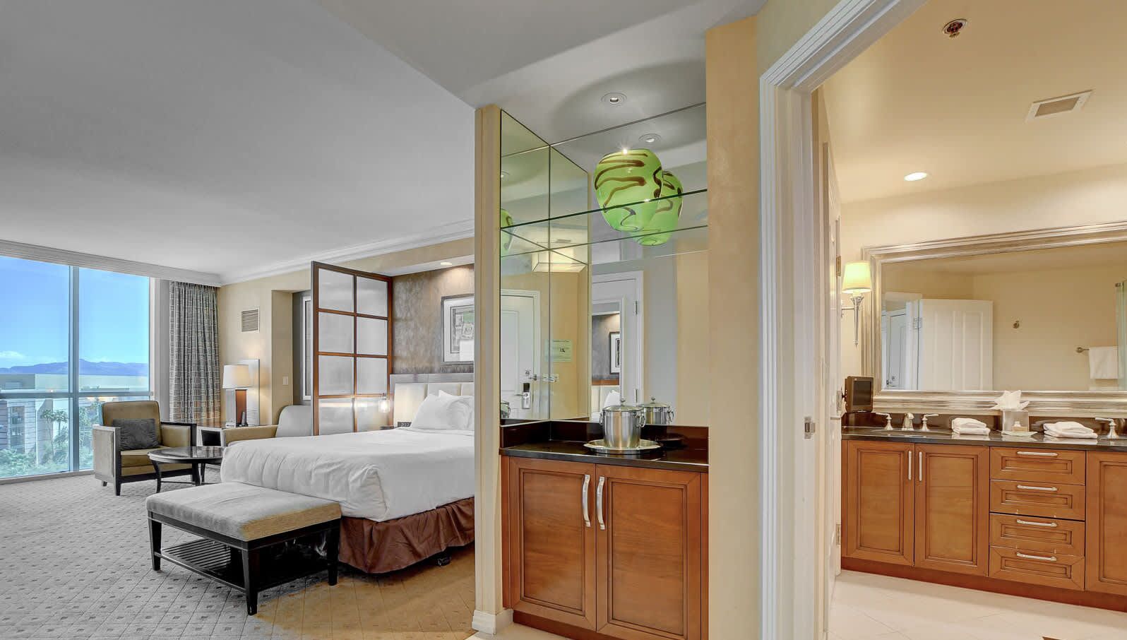 A hotel room with a large bed, a sitting area with a chair and table, a kitchenette, and a bathroom with a large mirror.