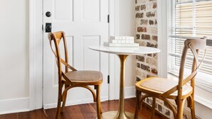 A room with a white door, a brick-patterned wall, and a window with blinds. There is a round table with two chairs. - Stylish 3BR | Sleeps 6 | Walk Everywhere (Charleston)