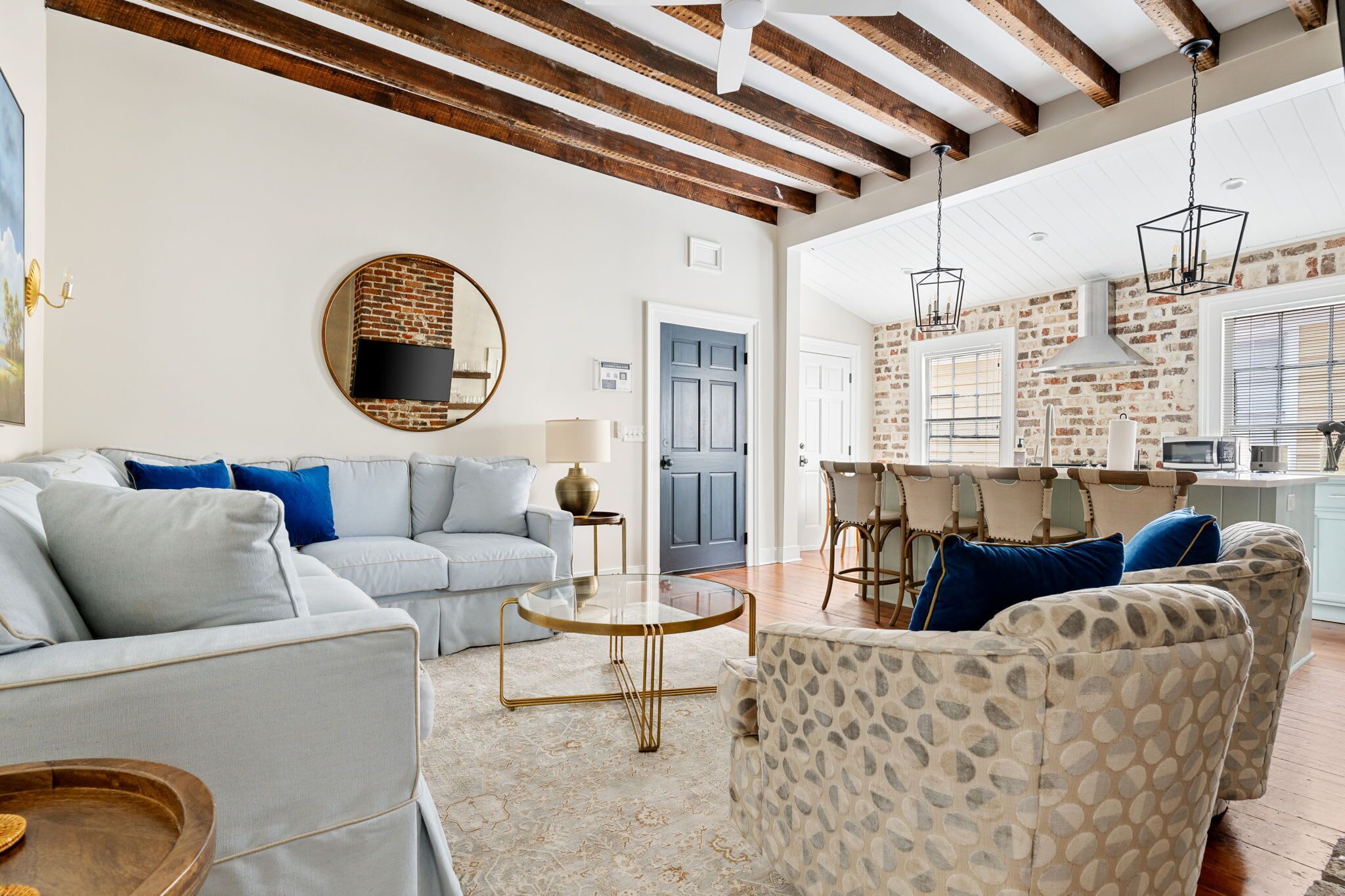 Amplia sala de estar con un sofá azul claro, sillones estampados, una mesa de centro redonda y una zona de cocina con paredes de ladrillo y luces colgantes.