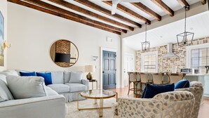 A spacious living room with a light blue sofa, patterned armchairs, a round coffee table, and a kitchen area with brick walls and hanging lights.