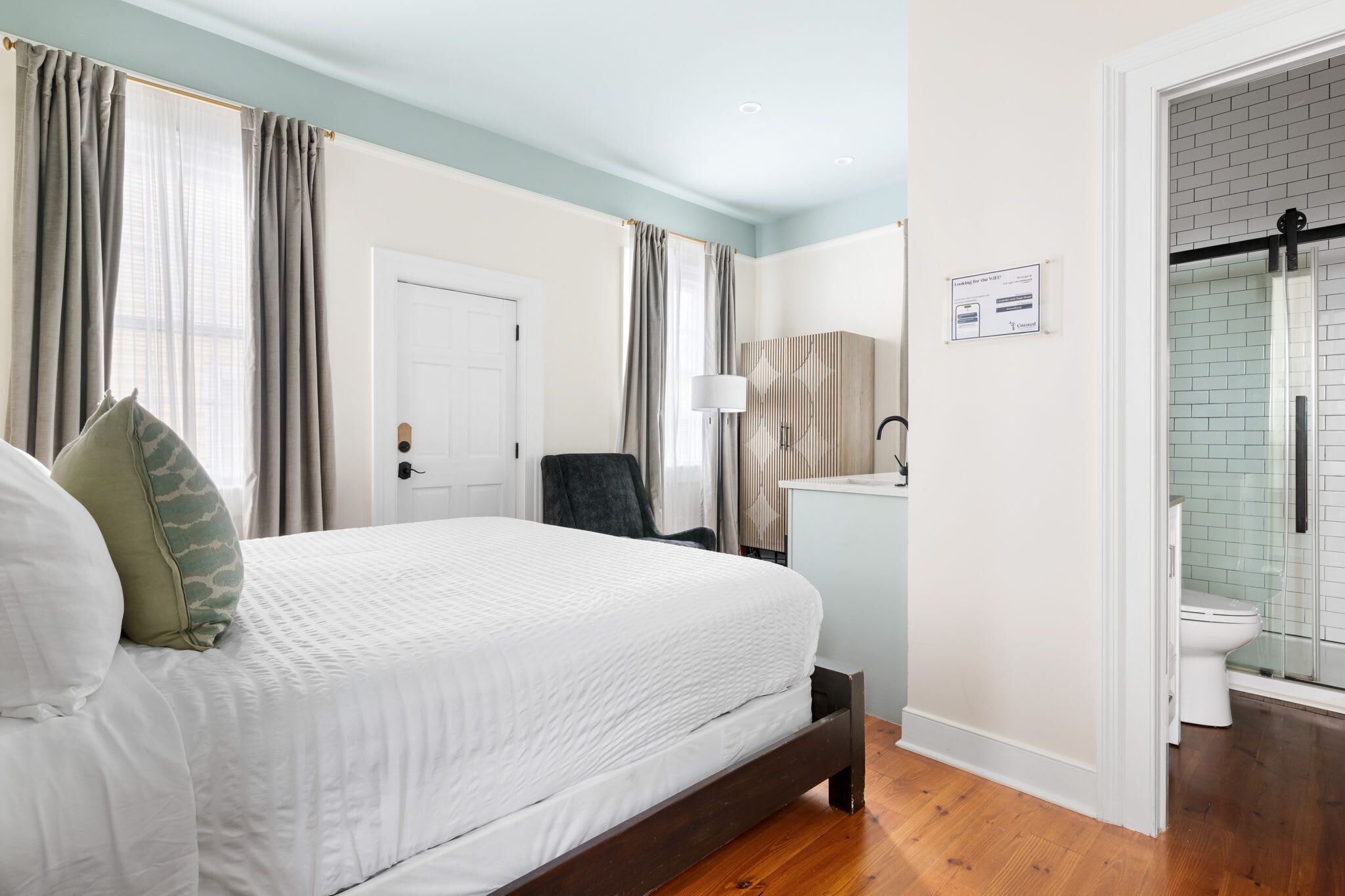 A bedroom with a bed, a chair, a desk, and a bathroom with a shower.
