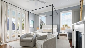 A bedroom with a four-poster bed, two armchairs, a nightstand, a fireplace, and large windows. - Downtown|3 Beds|Luxury Rental (Charleston)