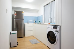 A compact kitchen with a stainless steel refrigerator, a washing machine, and a small trash bin.