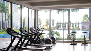 Une salle de sport équipée de tapis de course, d’elliptiques et d’un vélo d’appartement, de grandes fenêtres offrant une vue sur des palmiers et une piscine.