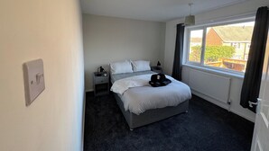 Ein Schlafzimmer mit einem Bett, einem Nachttisch, einer Lampe, einem Fenster mit Vorhängen und Blick auf Gebäude außerhalb.