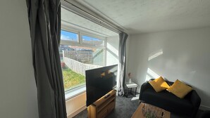 Ein modernes Wohnzimmer mit Flachbildfernseher, einem schwarzen Sofa mit gelben Kissen, einem Holztisch und Blick auf einen eingezäunten Garten durch große Fenster.