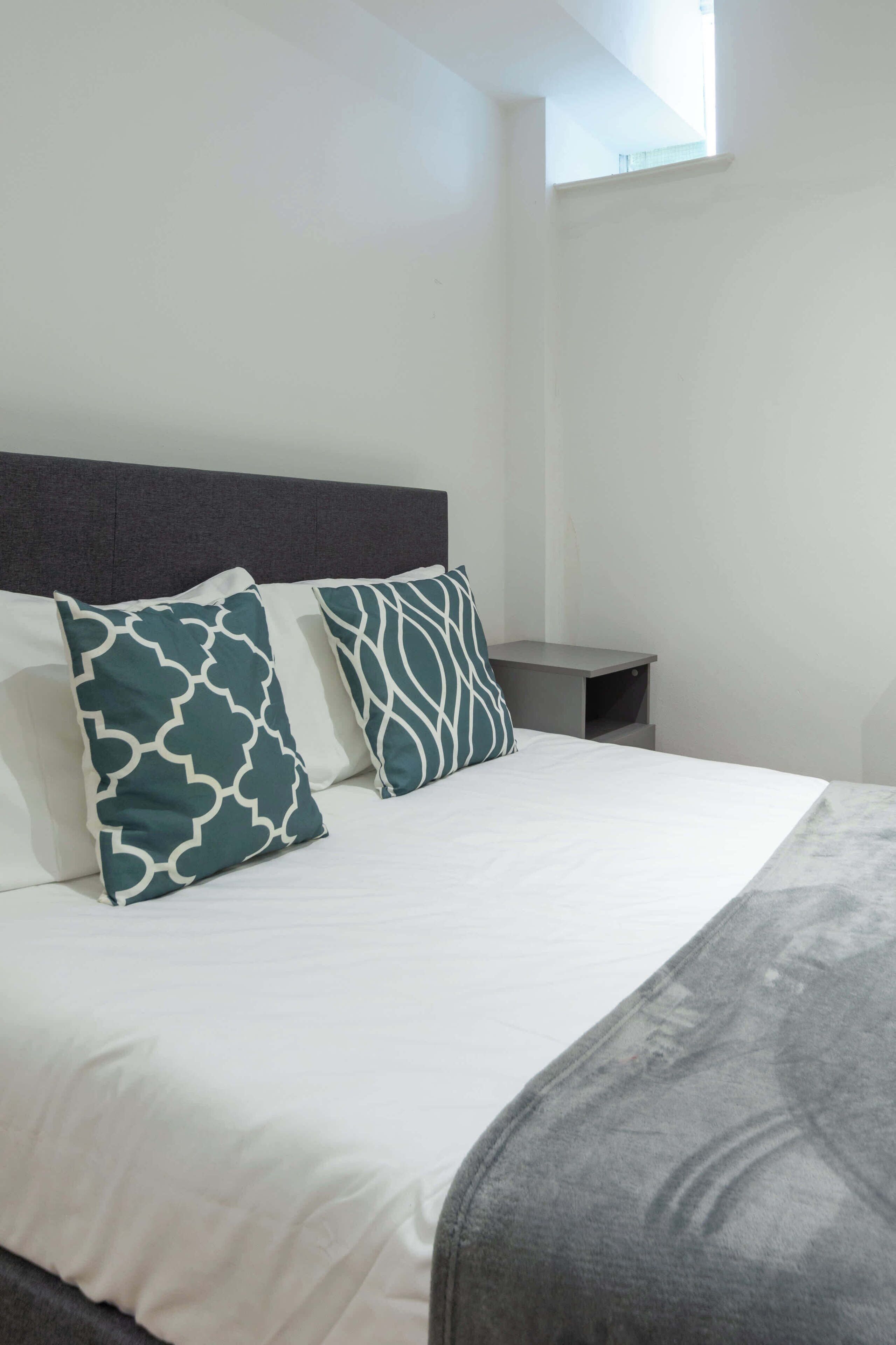 A neatly made bed with white bedding and two patterned pillows, a small bedside table, and a window with natural light.