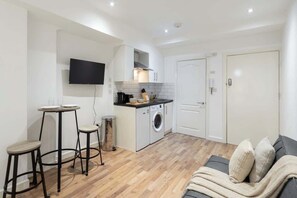 A compact living space with a kitchenette, a flat-screen TV mounted on the wall, a sofa with cushions, and a small table with stools. - Chic Shoreditch Studio with Wifi (London)