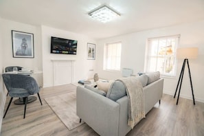 A modern living room with a grey sofa, a round coffee table, and a grey armchair. There is a flat-screen TV mounted on the wall, a lamp on a tripod, and a fireplace.