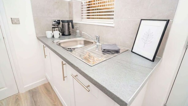 A compact kitchen with a sink, coffee maker, and a framed picture on the counter. - Stunning 1BR Cheltenham Suite Near Town Centre (Cheltenham)