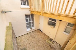 A small enclosed courtyard with a wooden balcony, two windows with shutters, and a gravel ground.