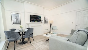 A modern living room with a grey sofa, a dining table with chairs, a flat-screen TV mounted above the fireplace, and framed pictures on the walls.