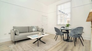 A modern living room with a grey sofa, a coffee table, and a dining table with chairs. There is a window with blinds, a door, and a small plant on the table. - The Cheltenham Suites - Stunning 2 Bed Apartment (Cheltenham)