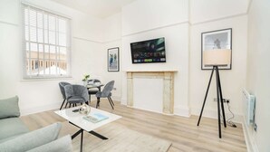 A modern living room with a fireplace, a television mounted on the wall, and a dining area with chairs and a table. - The Cheltenham Suites - Stunning 2 Bed Apartment (Cheltenham)