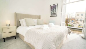 A modern bedroom with a large bed, a bedside table with a lamp, and a window with a view of buildings. - The Cheltenham Suites - Stunning 2 Bed Apartment (Cheltenham)