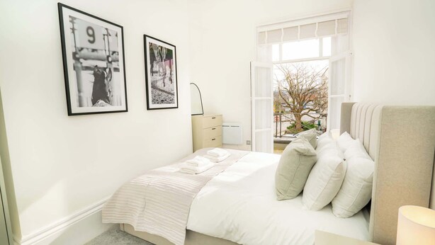 A bedroom with a bed, a dresser, a window with a view, and two framed pictures on the wall. - The Cheltenham Suites - Stunning 2 Bed Apartment (Cheltenham)