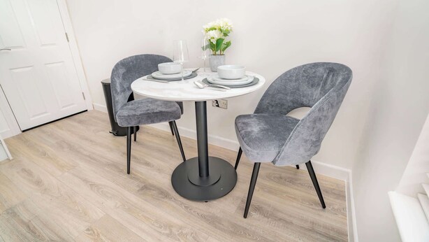 A dining area with a round table, two grey upholstered chairs, a vase with white flowers, and a white door in the background.