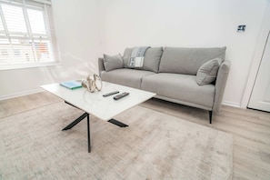 Uma sala de estar moderna com um sofá cinzento, uma mesa de centro branca e um tapete pequeno.