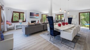 Ein modernes Wohnzimmer mit einer Couch, einem Esstisch und Stühlen. Große Fenster mit Vorhängen, ein Gemälde an der Wand und ein Blick auf Grünflächen im Freien.