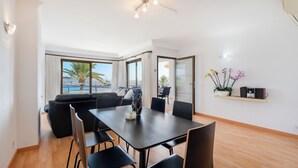 A modern living room with a black sofa, a dining table with chairs, a coffee table, a potted plant, and a view of palm trees and a cityscape through large windows. - Nitas Apartment (Pollença)