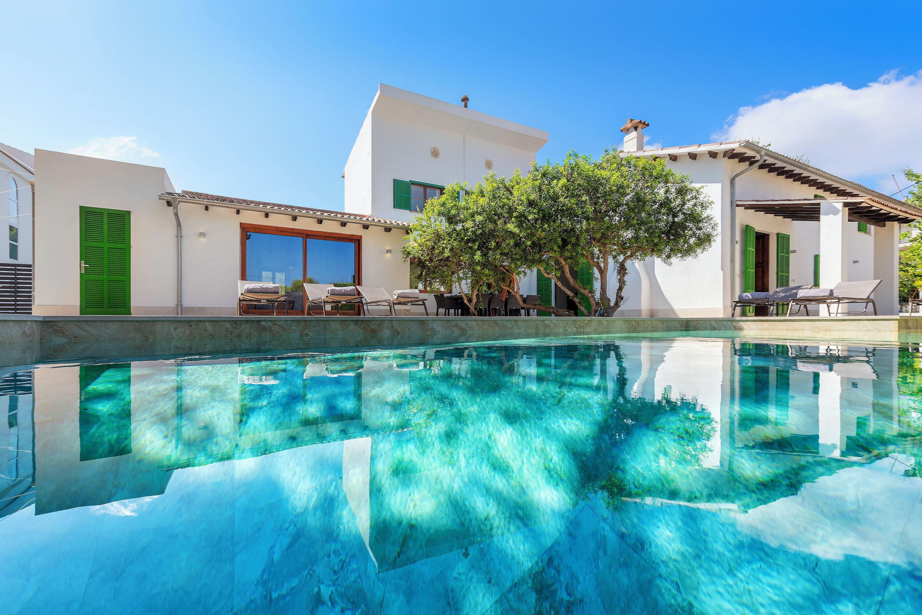 Un área de piscina con agua cristalina, un edificio blanco con contraventanas verdes y un árbol en primer plano.
