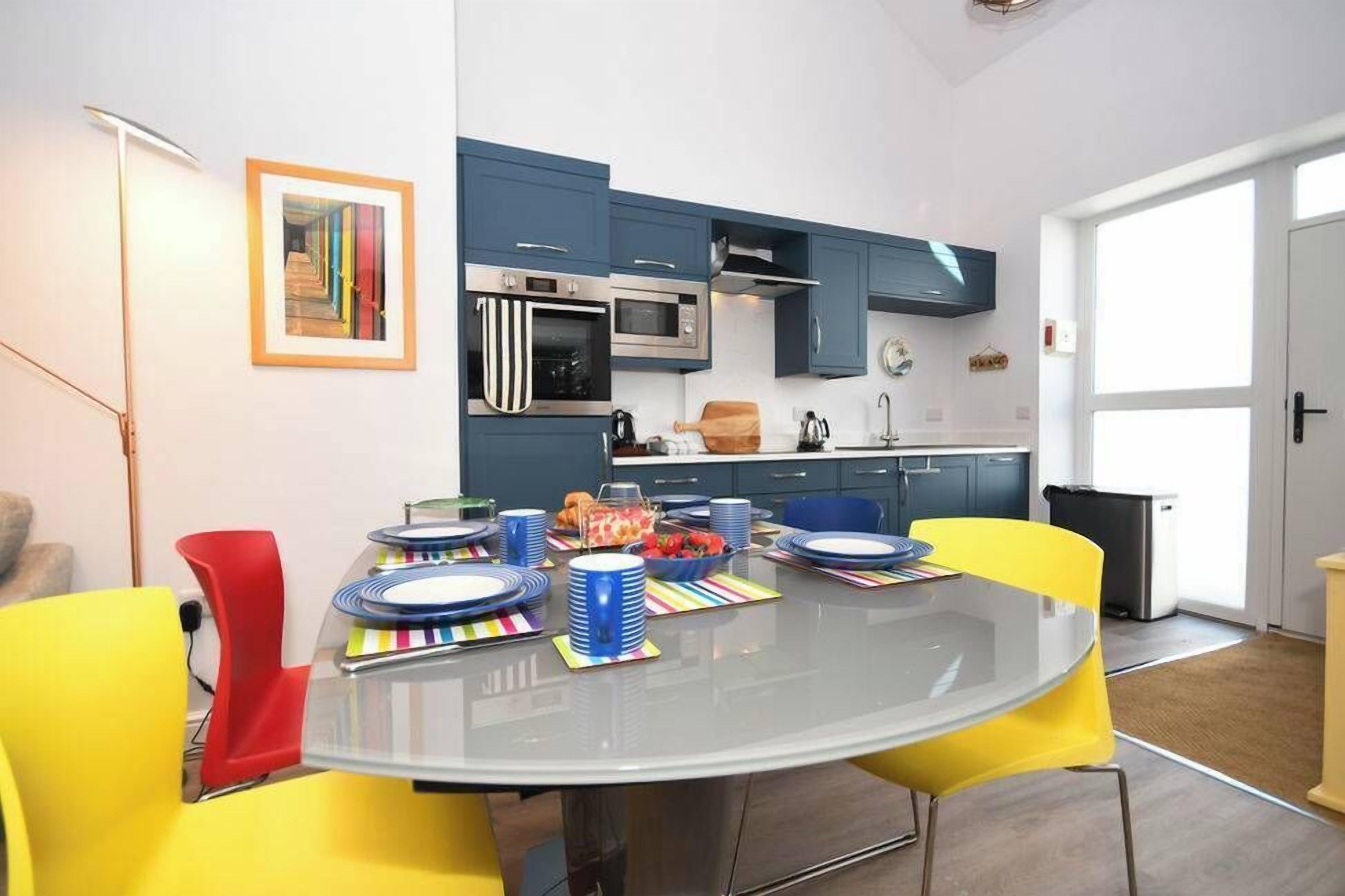 A modern kitchen with blue cabinets, a microwave, and a stove. There is a dining table with yellow chairs and a glass top.