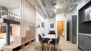 Un espace de vie moderne et compact comprenant un lit superposé, un coin repas avec une table et des chaises en bois, un coin cuisine avec un réfrigérateur et un petit coin salon avec un canapé.