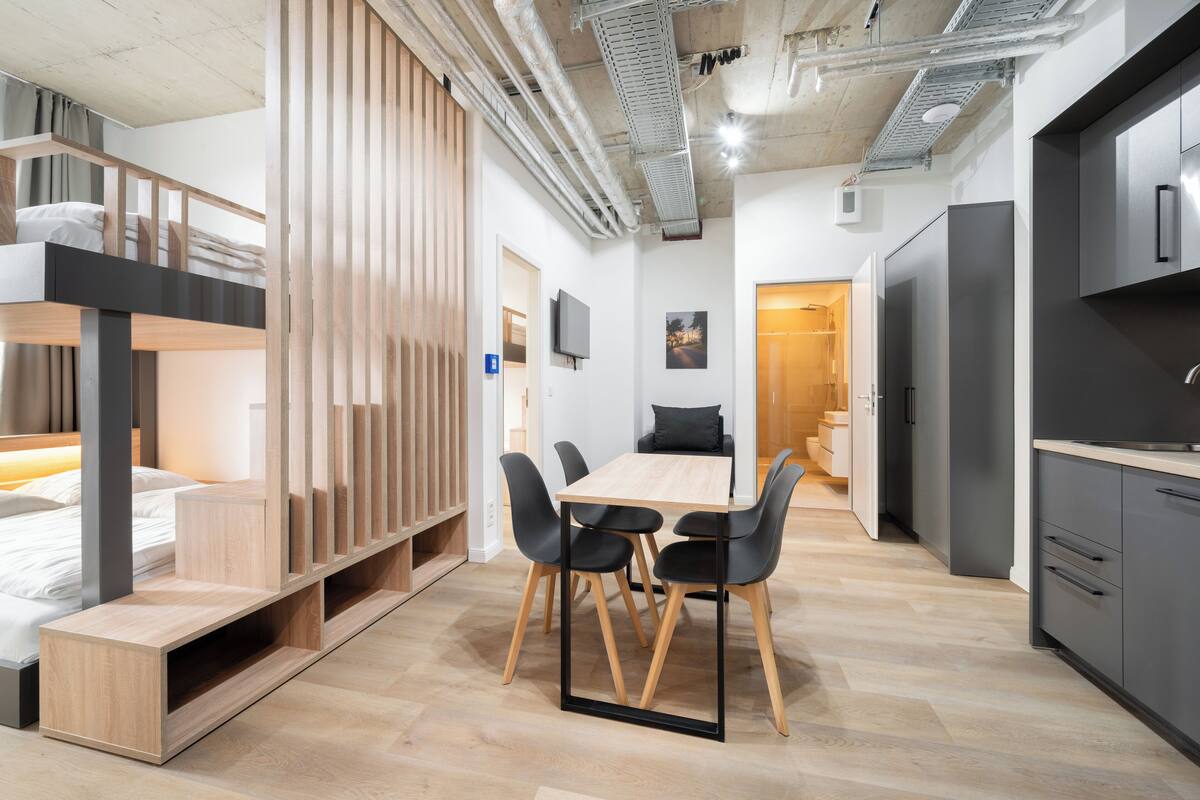 Ein moderner, kompakter Wohnraum mit Etagenbett, Essbereich mit Holztisch und Stühlen, Küchenzeile mit Kühlschrank und einer kleinen Sitzecke mit einer Couch.