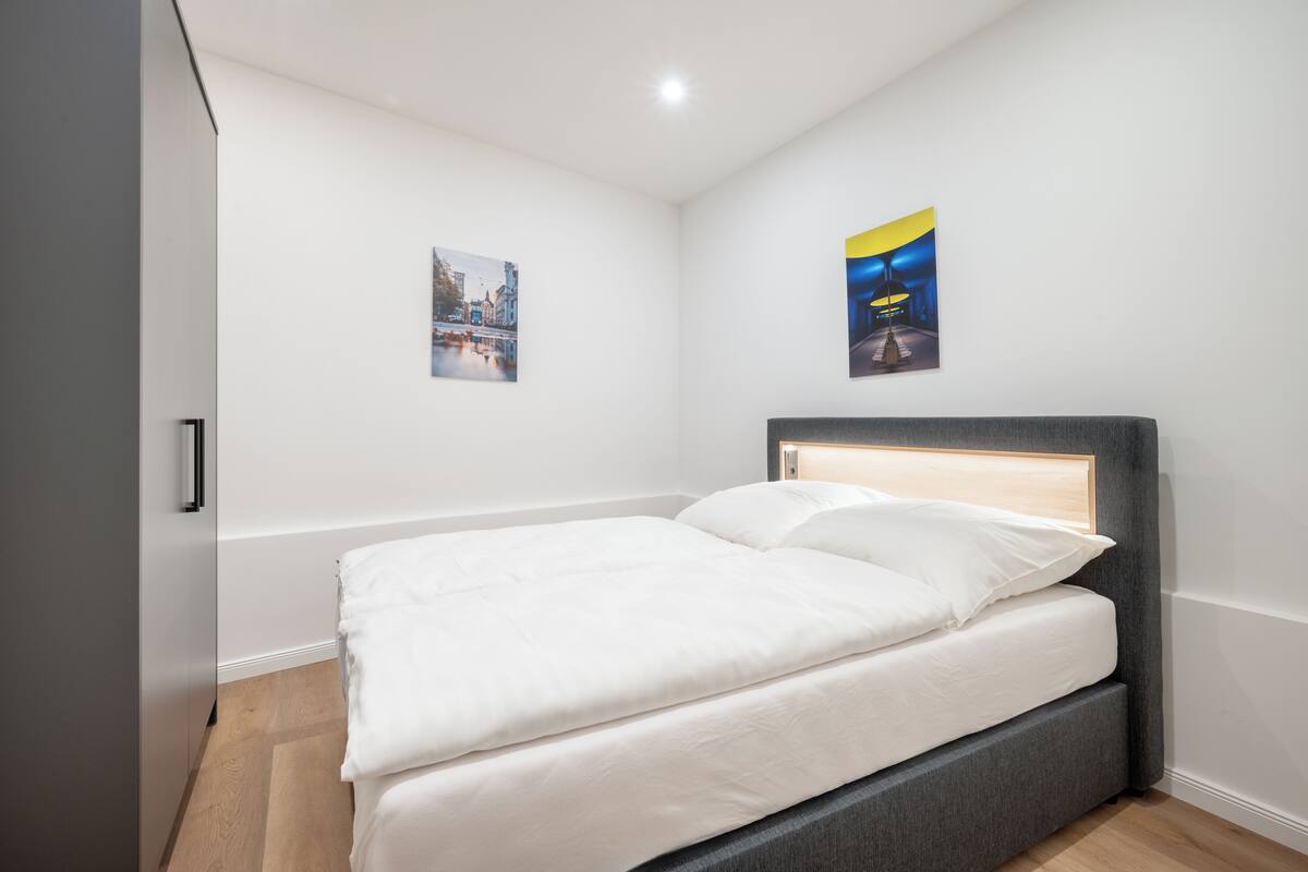 Ein ordentlich bezogenes Bett in einem modernen Zimmer mit grauem Kopfteil, weißen Wänden und Holzboden.