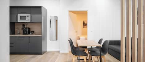 A modern kitchen with grey cabinets, a microwave, and a coffee maker. There is a dining area with a wooden table and black chairs.