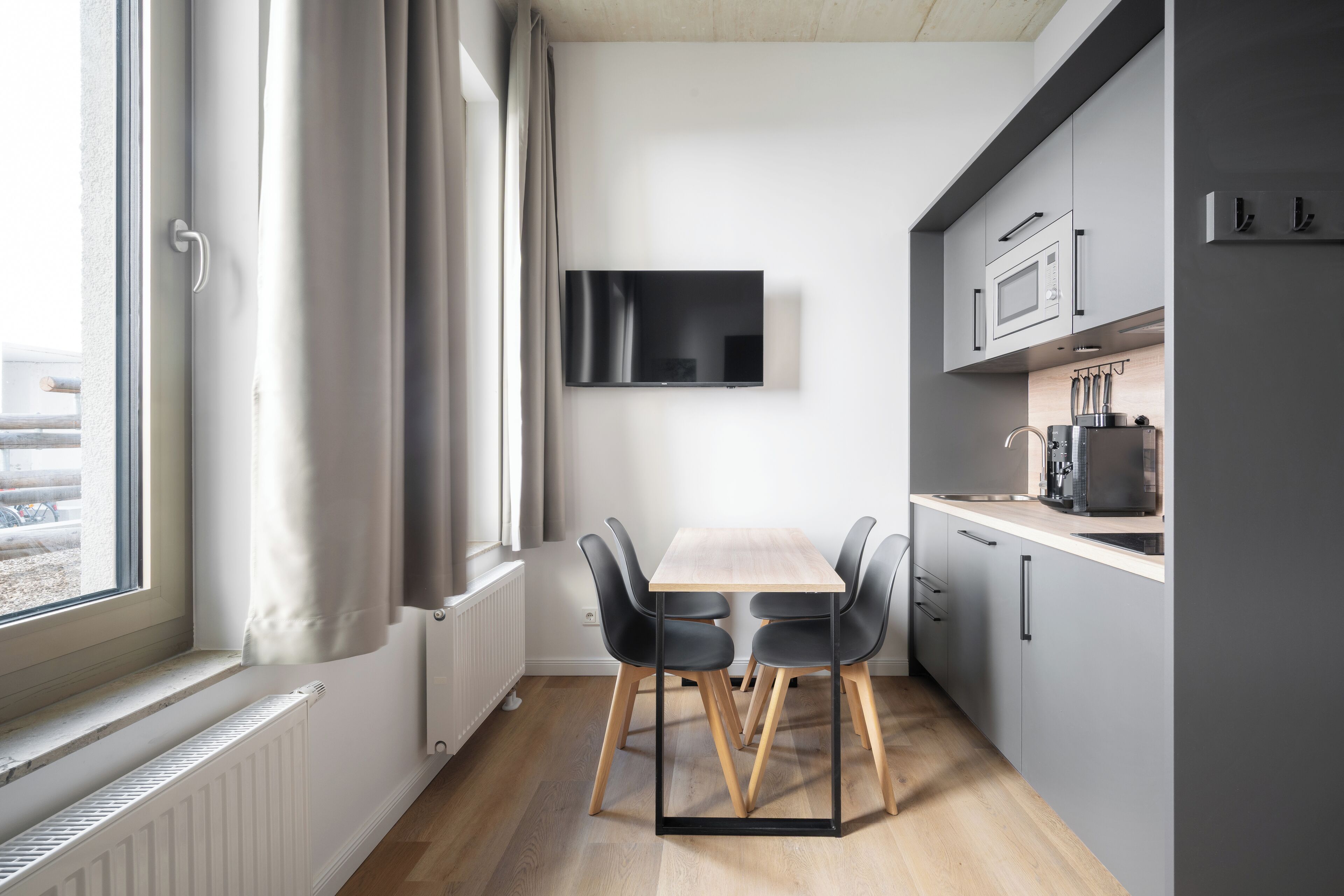 Una cocina moderna con una mesa y sillas de madera, un televisor de pantalla plana sobre la encimera, y un ventanal con cortinas.