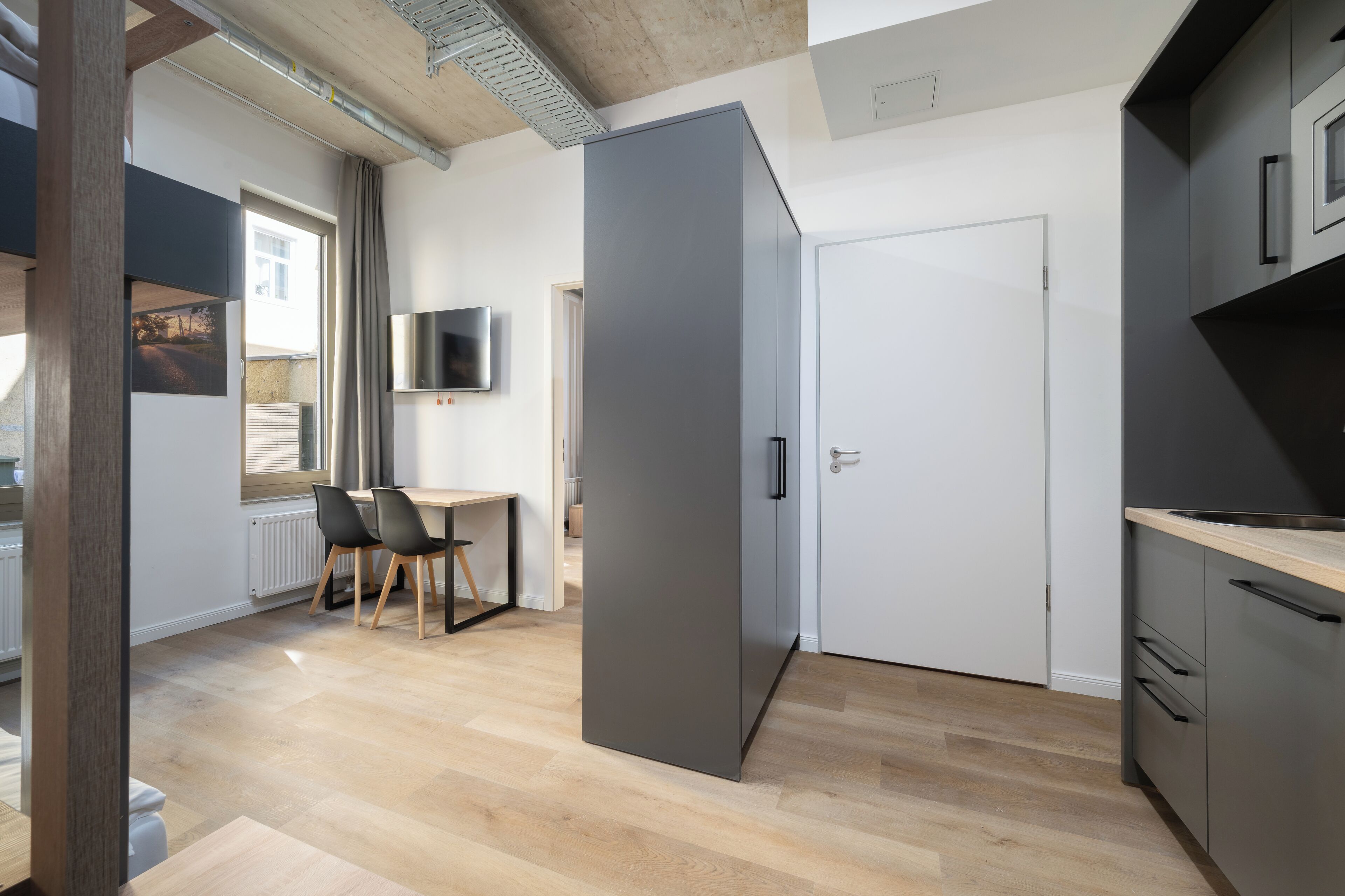 A modern kitchen with grey cabinets, a wooden dining table with chairs, and a TV mounted on the wall.