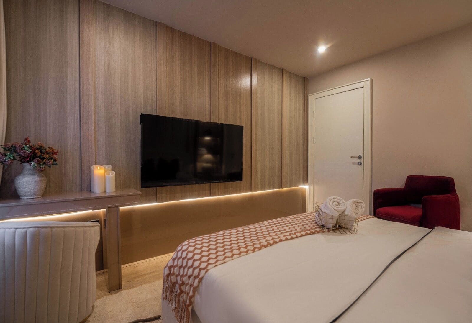Una habitación de hotel moderna con una cama grande, un televisor montado en la pared, una silla roja y una mesita de noche con velas.