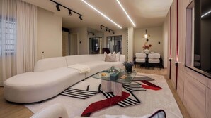 A modern living room with a curved white sofa, a glass coffee table, and a dining area with white chairs. - Libertini Apartments by FreedomWay (Lekki)