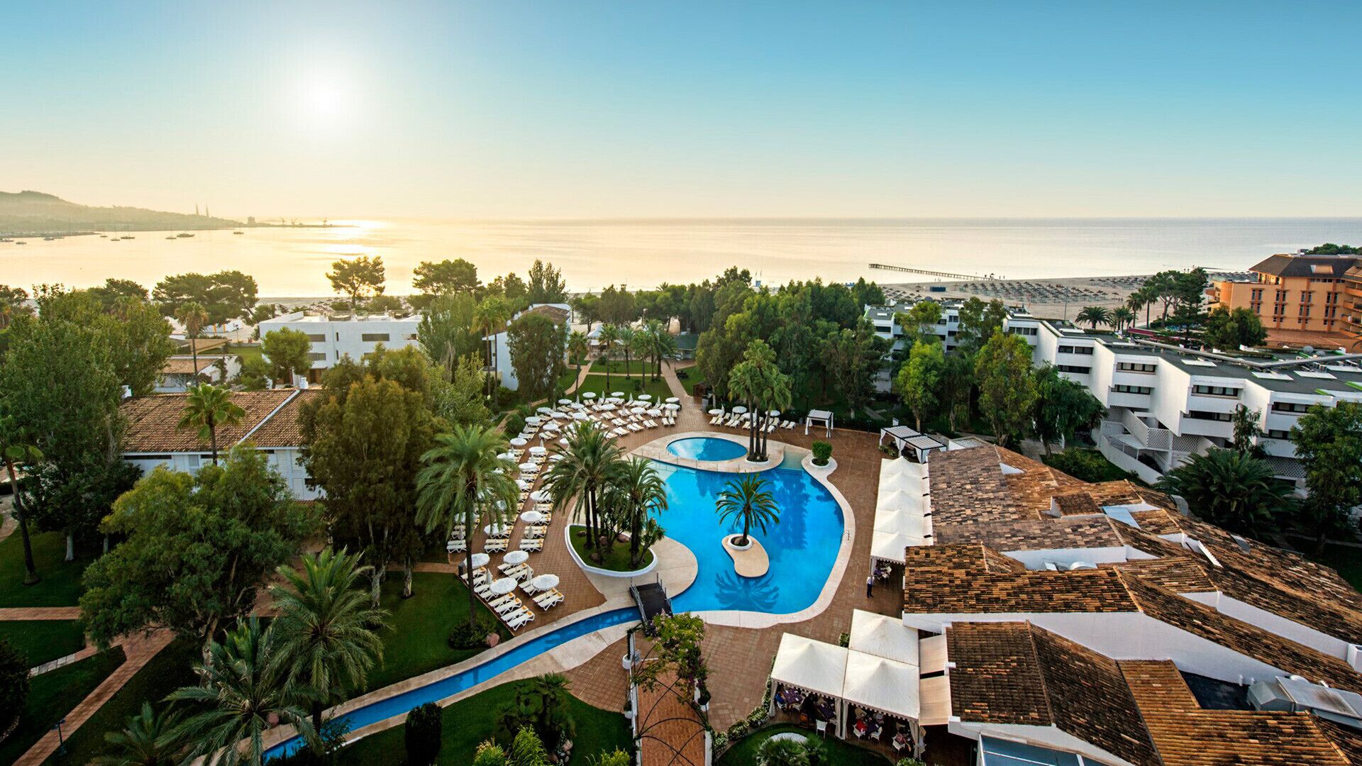 Ovenudblik over et resort med en central swimmingpool, liggestole og udsigt til havet.