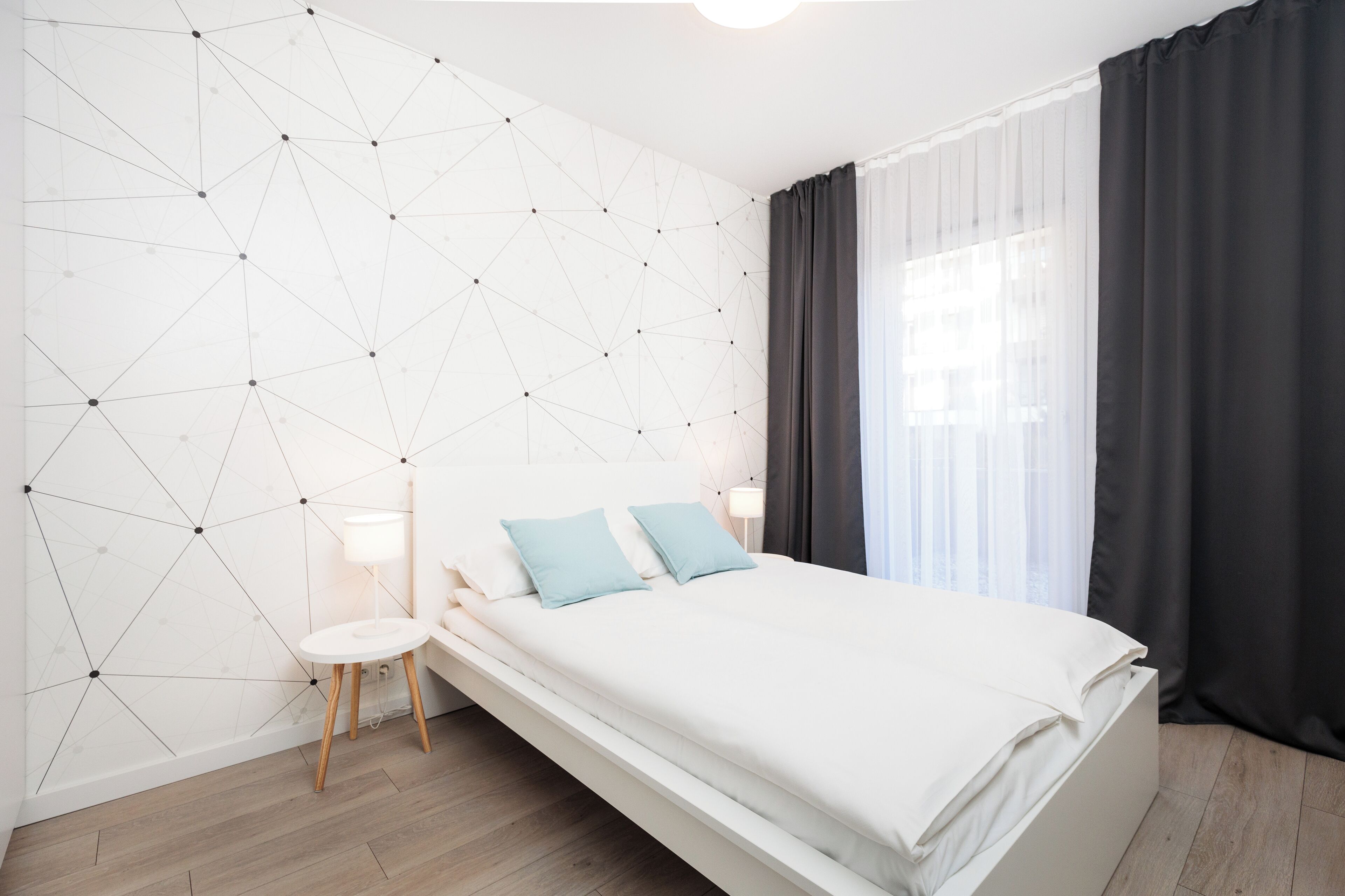 Une chambre moderne avec un mur orné d’un motif géométrique, un lit blanc avec deux oreillers, une table de chevet avec une lampe et une fenêtre avec des rideaux foncés.