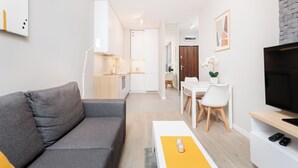 A modern living room with a grey sofa, a white coffee table with a yellow placemat, a flat-screen TV on a wooden stand, and a dining area with a white table and chairs. - City of the Kings-Milford Sound (Kraków)